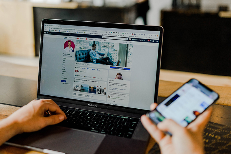 laptop screen flashes Facebook feed while a man holding a cellphone