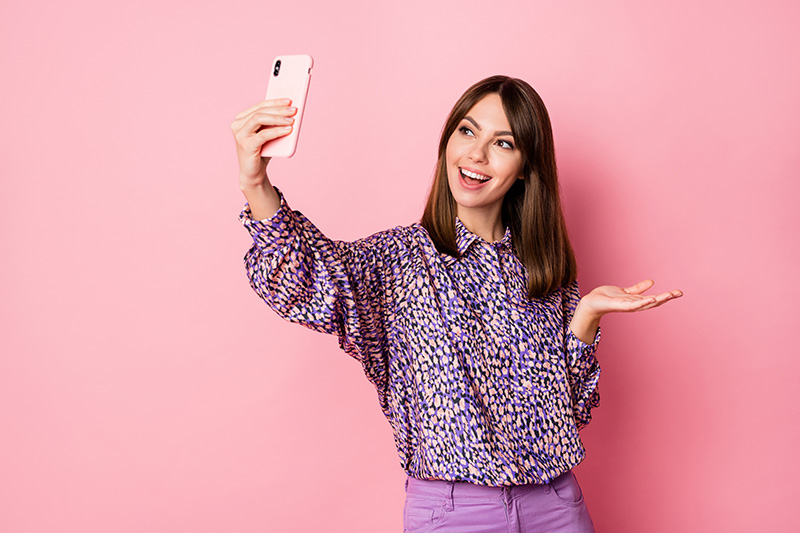 woman taking selfie holding phone in one hand isolated on pastel pink colored background with blank space