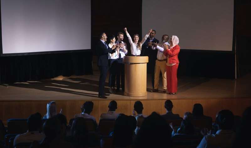 Happy businesswoman standing at the stage of the auditorium with colleagues in front of audience